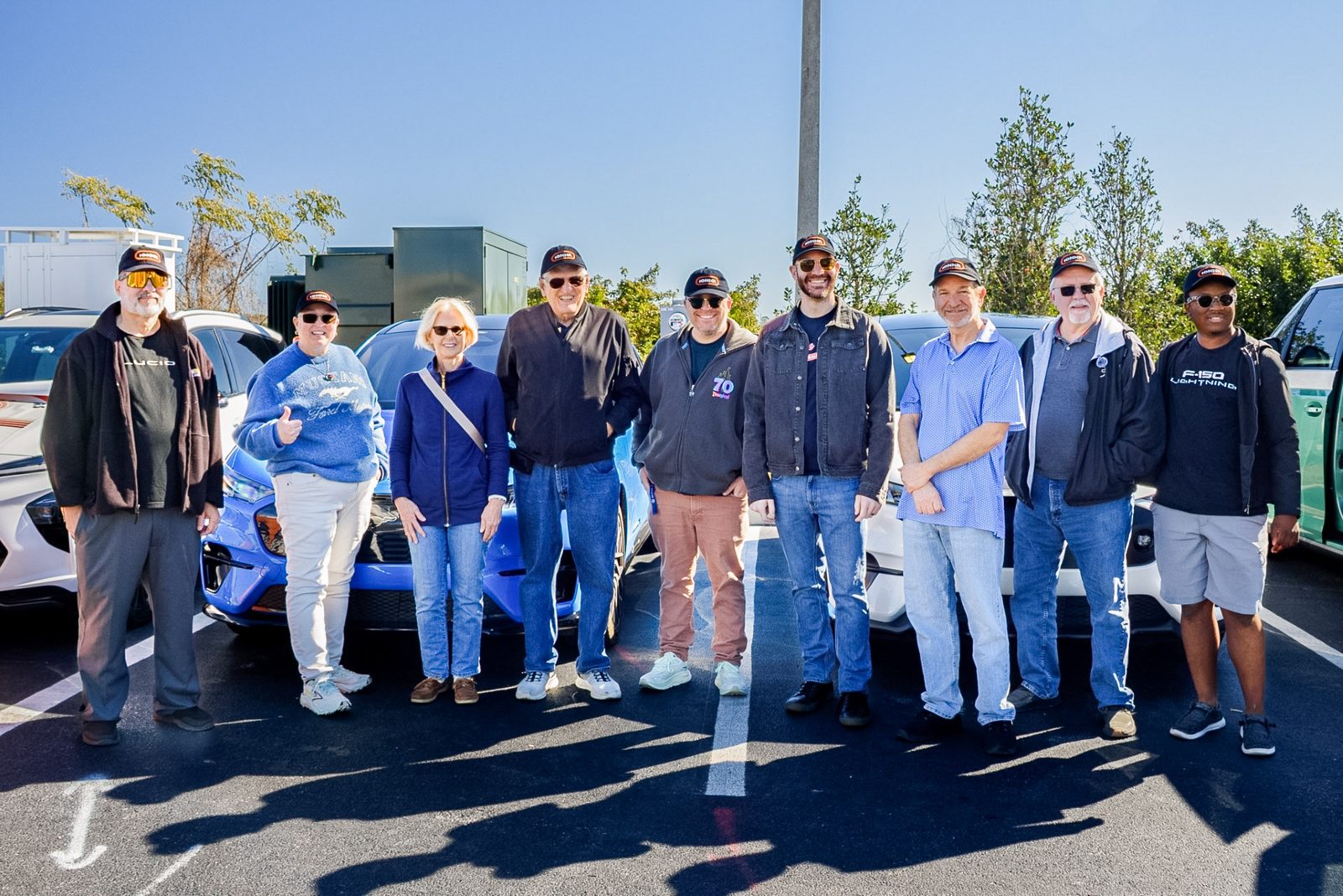 Nine drivers in IONNA caps lined up in front of a blue Mustang Mach-E at a get-together