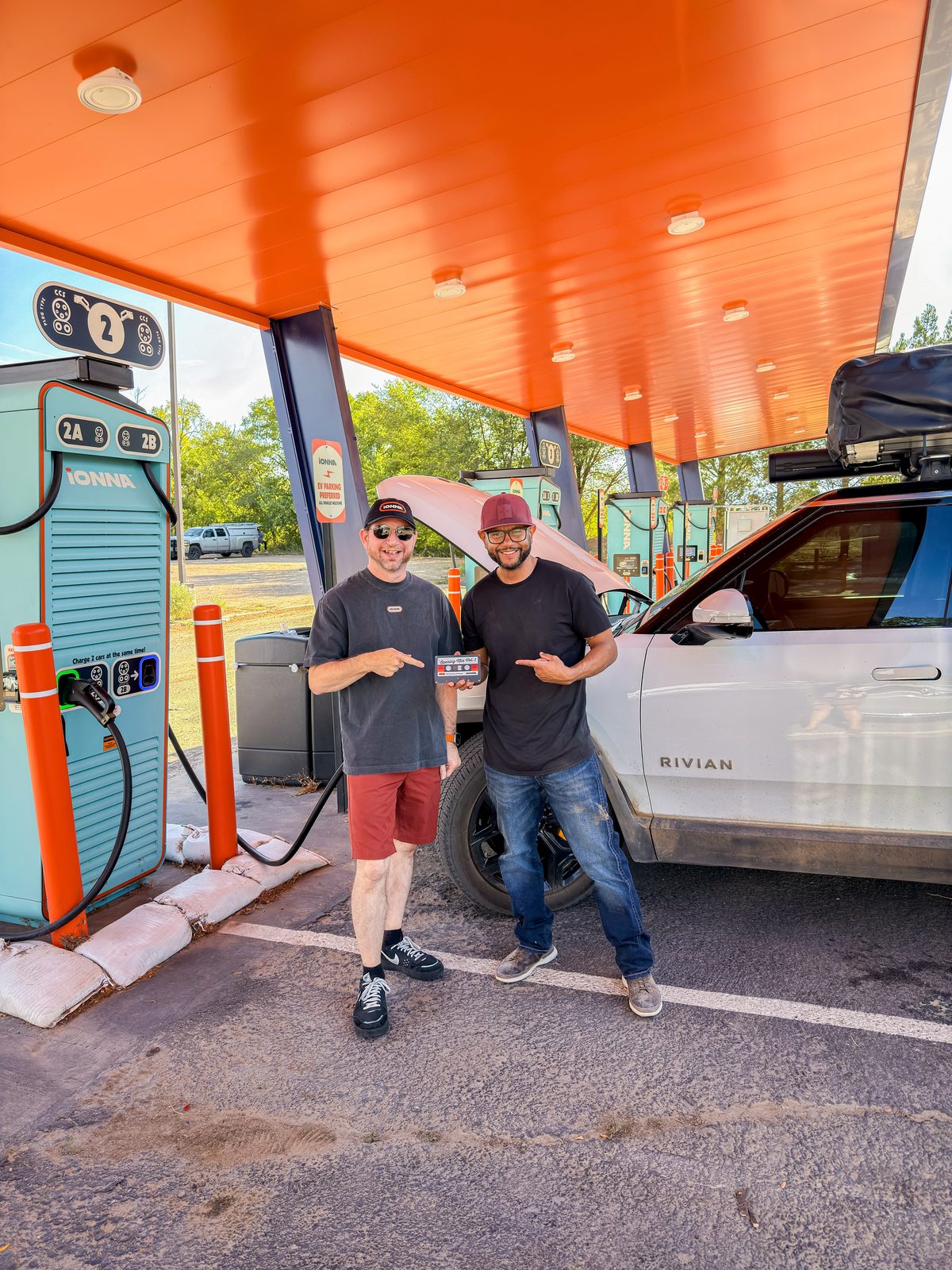 Two Station Hands under the orange IONNA canopy holding up a charging mixtape, with a white Rivian parked alongside